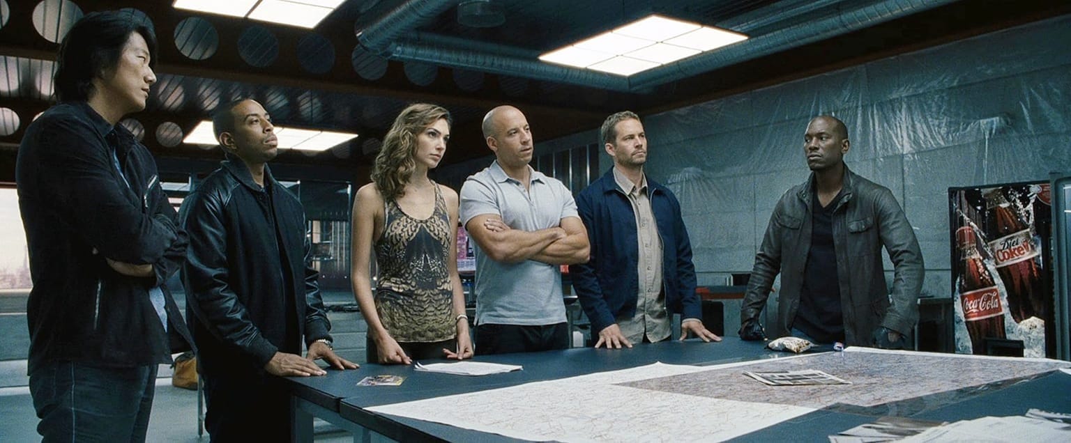 The squad plans a job at a big table with maps and a Coke machine