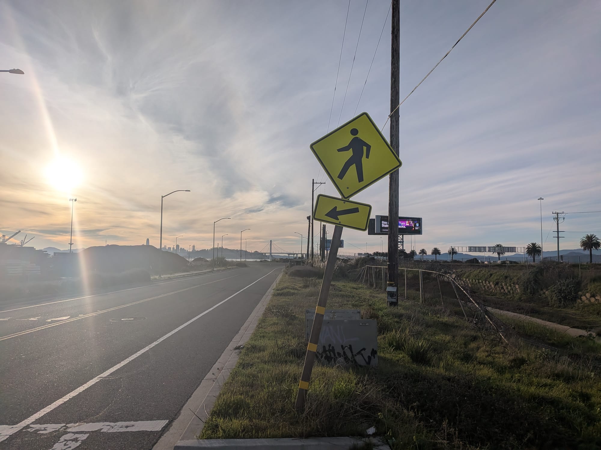 The West Oakland landscape, the West Bay skyline in the distance, with a sun flare and a pedestrian crossing sign in the foreground.
